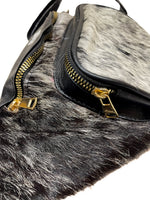 Close-up of black cow hair fanny pack showing front zippered pocket with genuine cowhide and brown leather trim. 