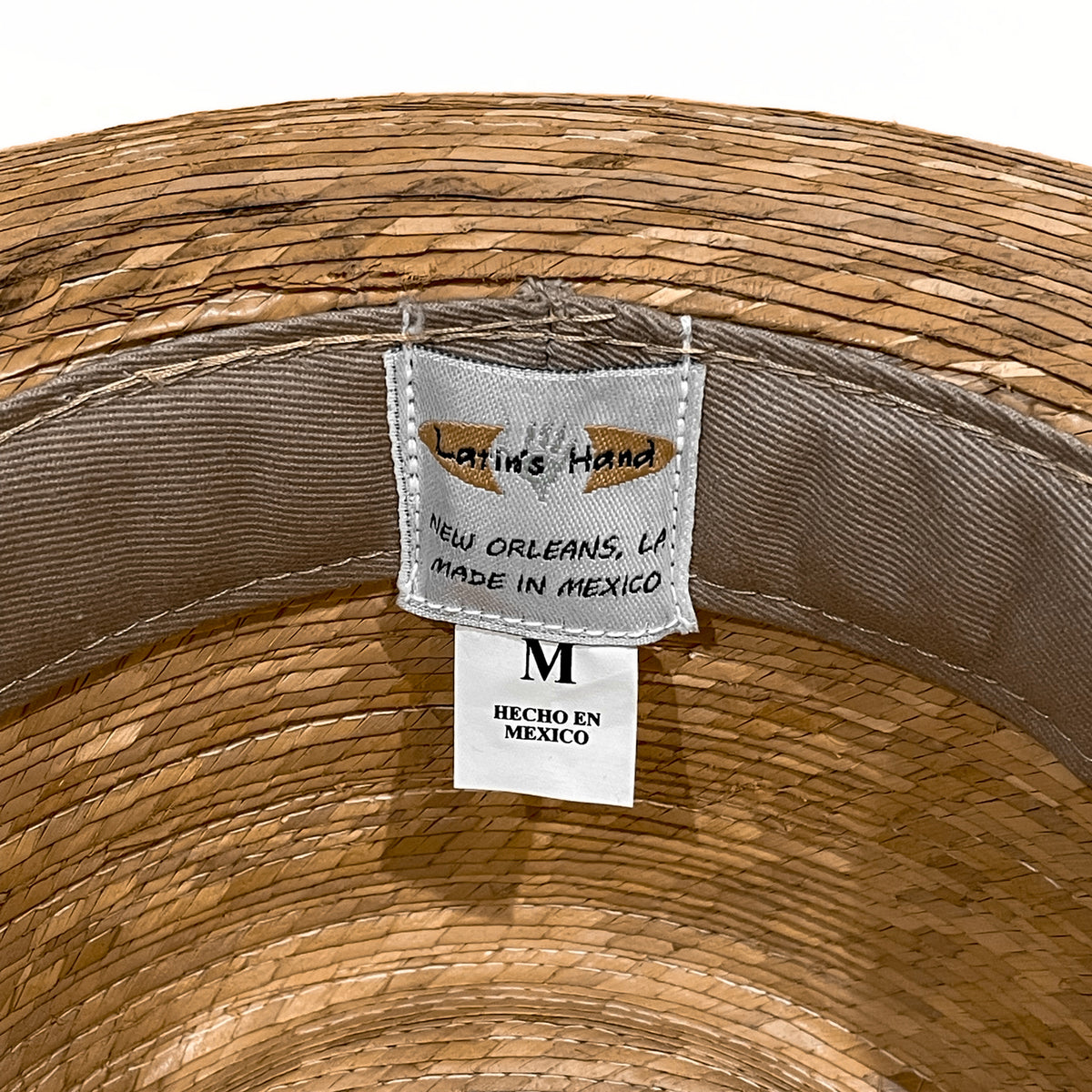 Close-up of the inside of a woven straw hat showing a sewn-in label that reads 'Latinos Hand' and 'New Orleans, LA Made in Mexico' with a size M tag below. 